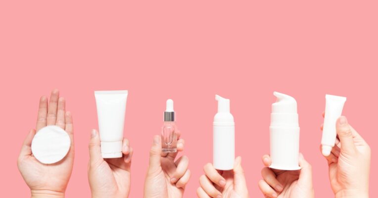 A lineup of unlabeled beauty products in various containers, held up by six hands against a pink background.