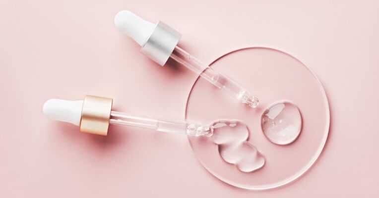 Two liquid pipette droppers dispense a clear serum into a clear dish against a pastel pink background.