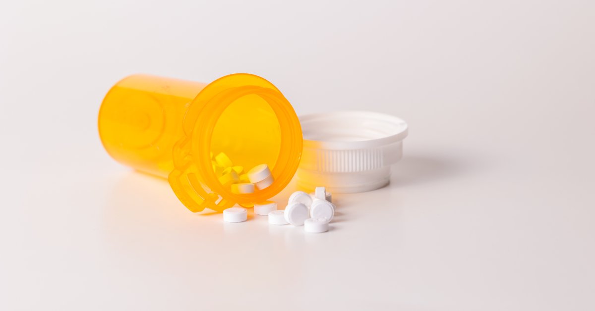 An orange labelless prescription pill bottle lies on a white background with white pills spilling out of it.