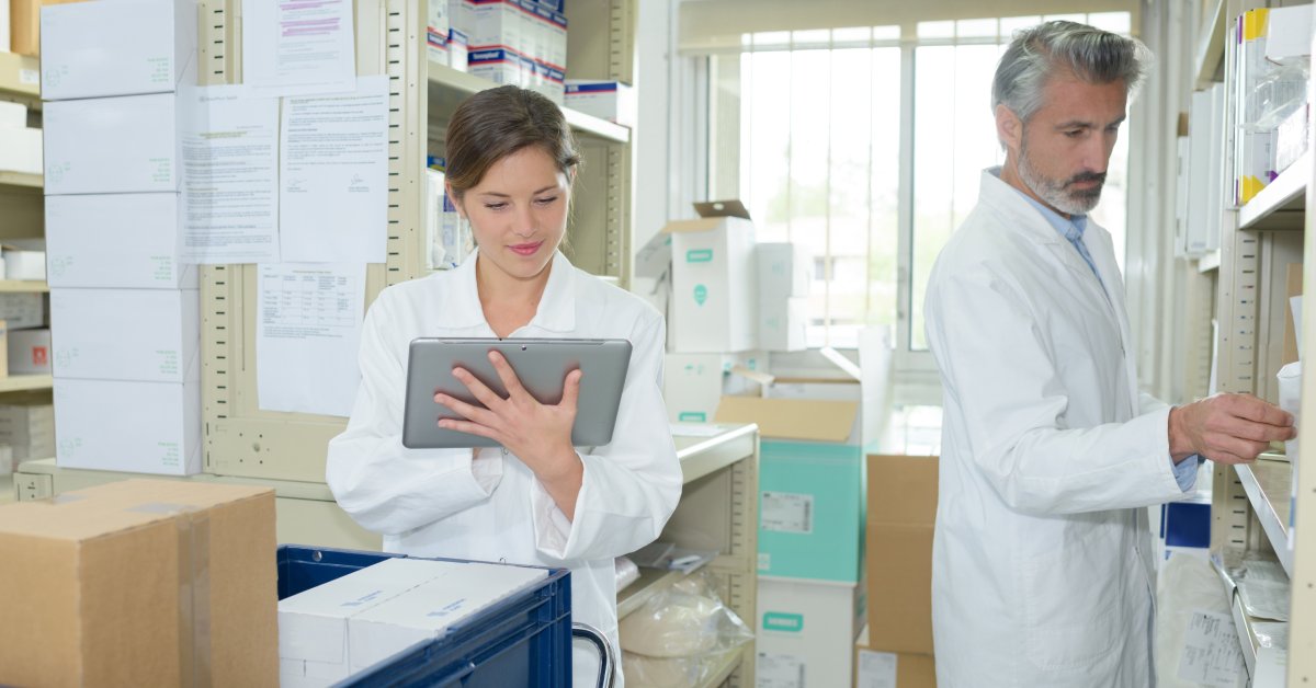 A male and a female pharmacy worker, each dressed in lab coats, work in a storage room. The woman holds a tablet.