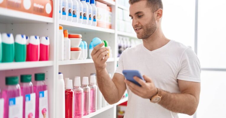 A man in a white t-shirt stands in a health and beauty aisle, holding a deodorant bottle in his hand.